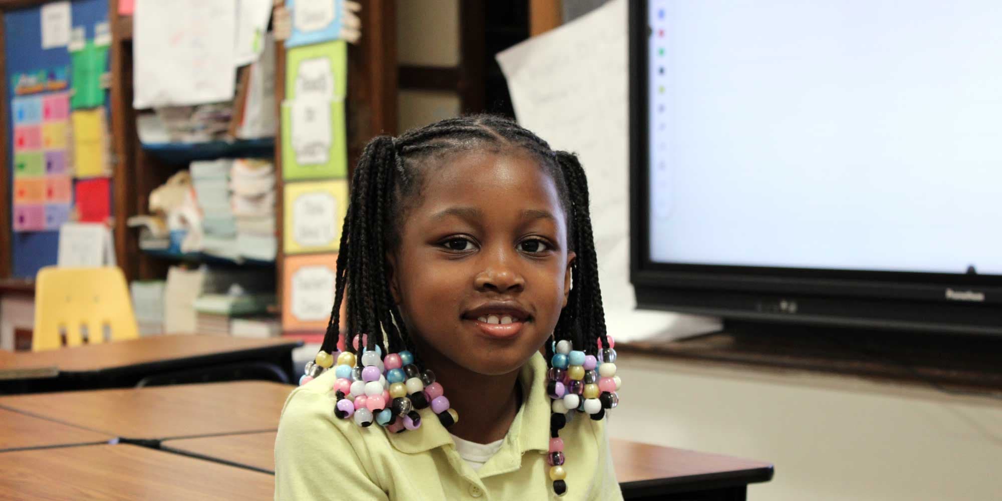 Smiling elementary student with beaded braids at a desk in front of a classroom display screen.