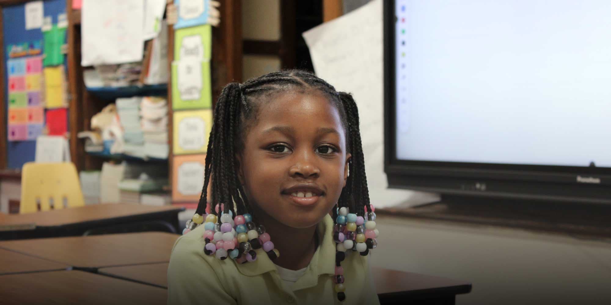 Smiling elementary student with beaded braids sitting at a desk in a Barber Prep classroom.