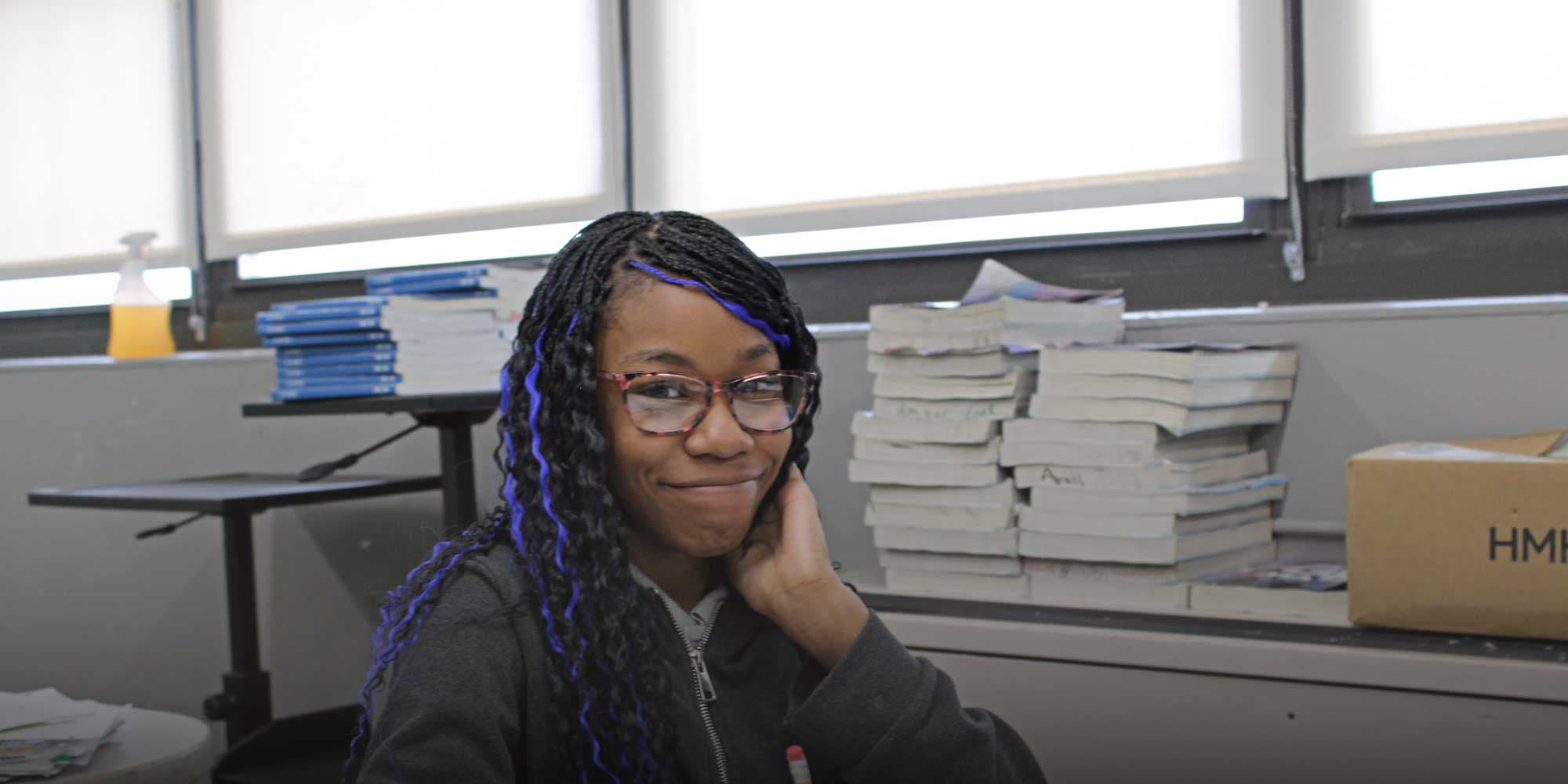Smiling middle school student with glasses seated at a desk in a classroom.