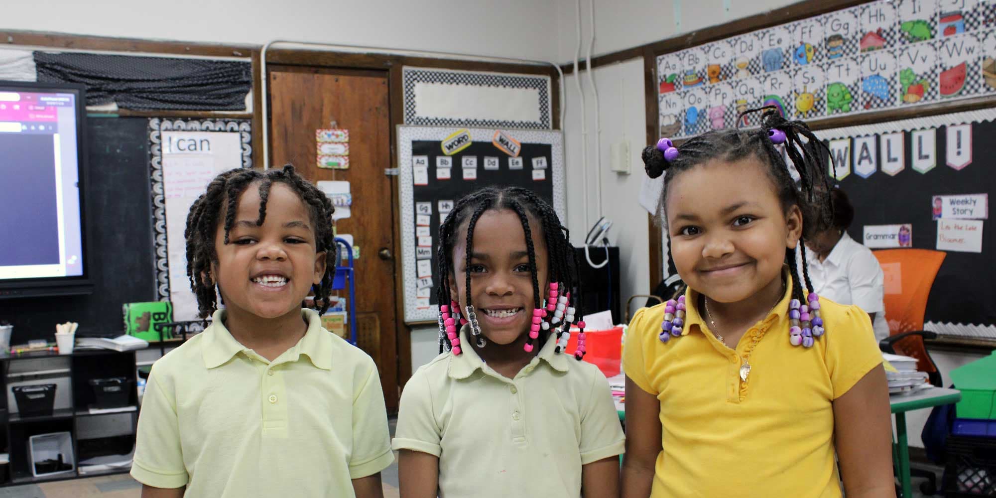 Three smiling young students in school uniforms standing in a classroom.