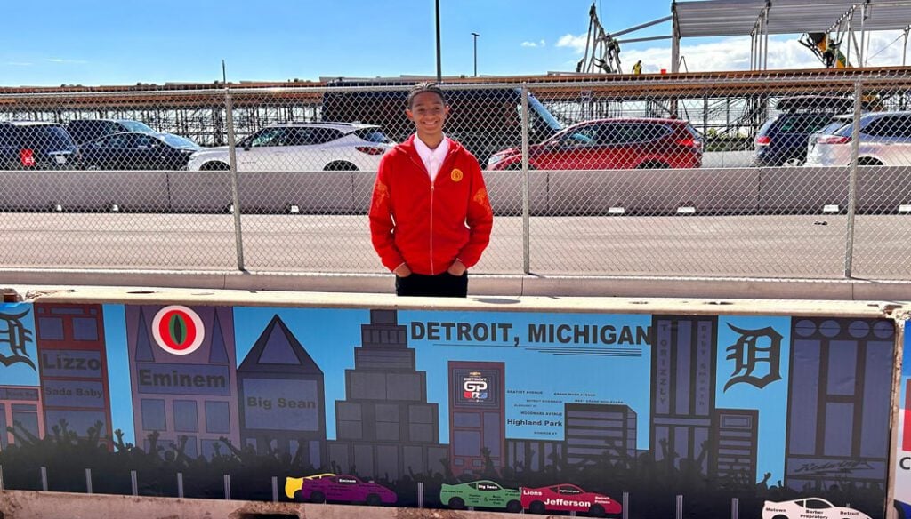 Adult in red jacket in front of a Detroit cityscape mural with race cars.