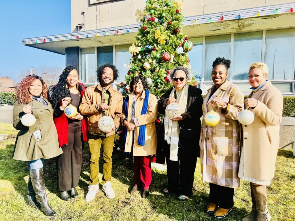 Seven adults hold ornaments in front of a Christmas tree.