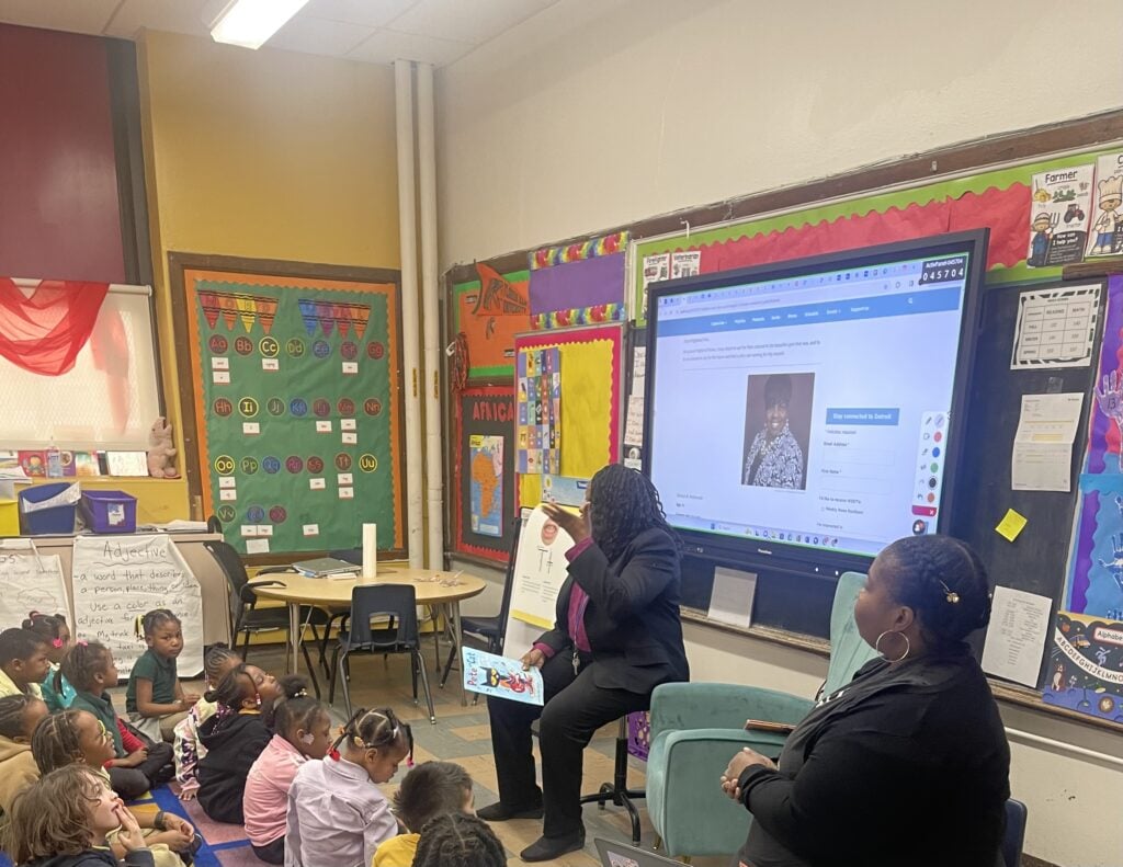 Adults reading to children in a classroom with a large screen.