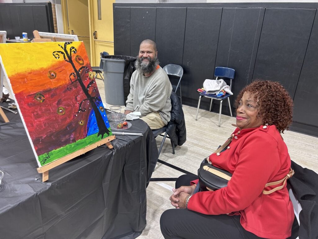 Adults seated with a colorful painting on an easel.