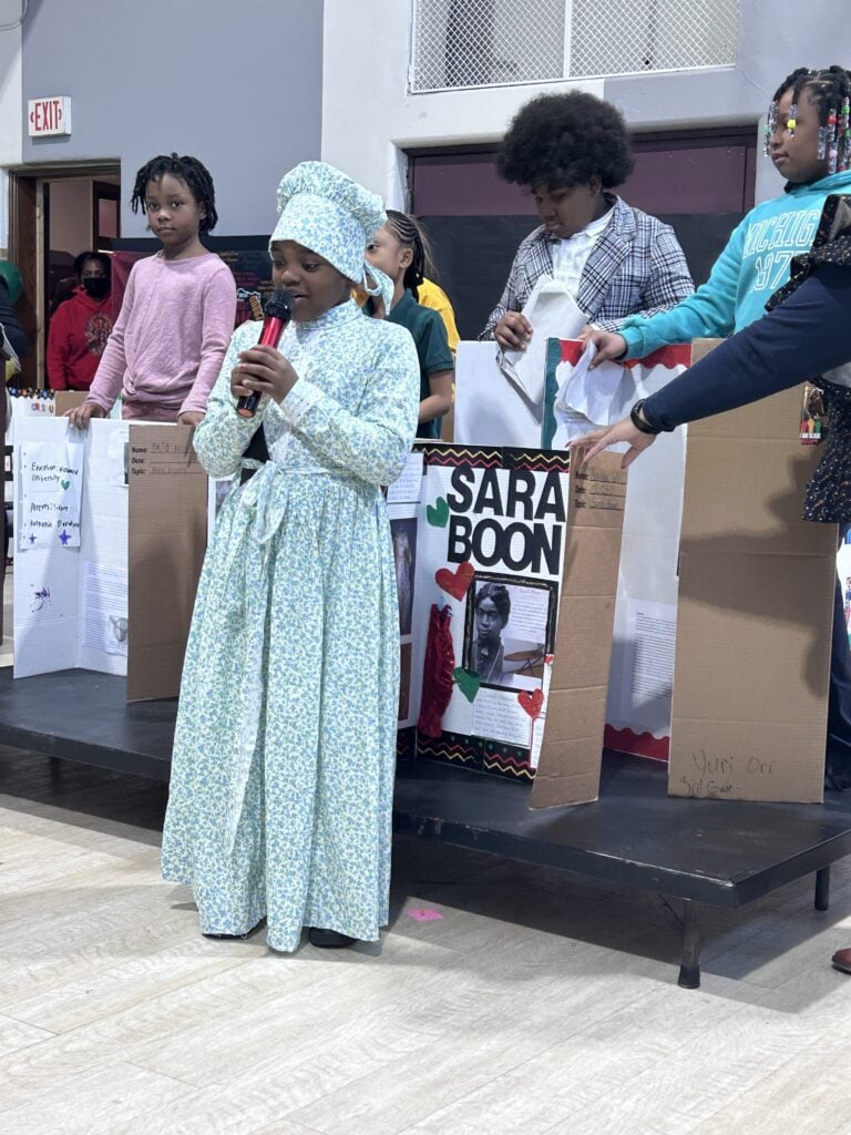 Child in historical costume speaks into microphone at school event.