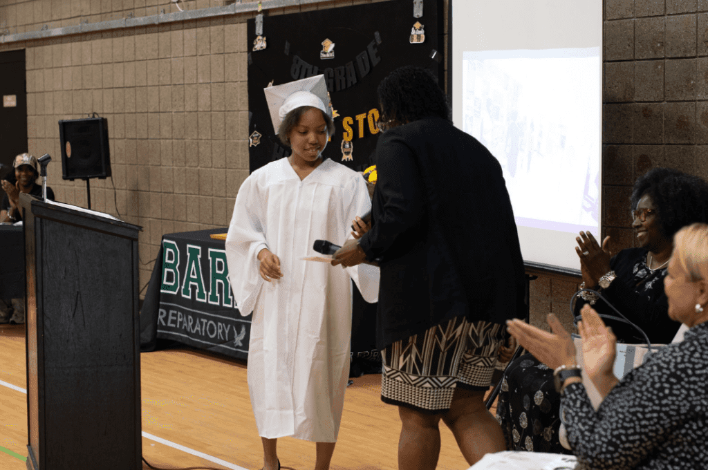 Child in white graduation gown receives award from adult.
