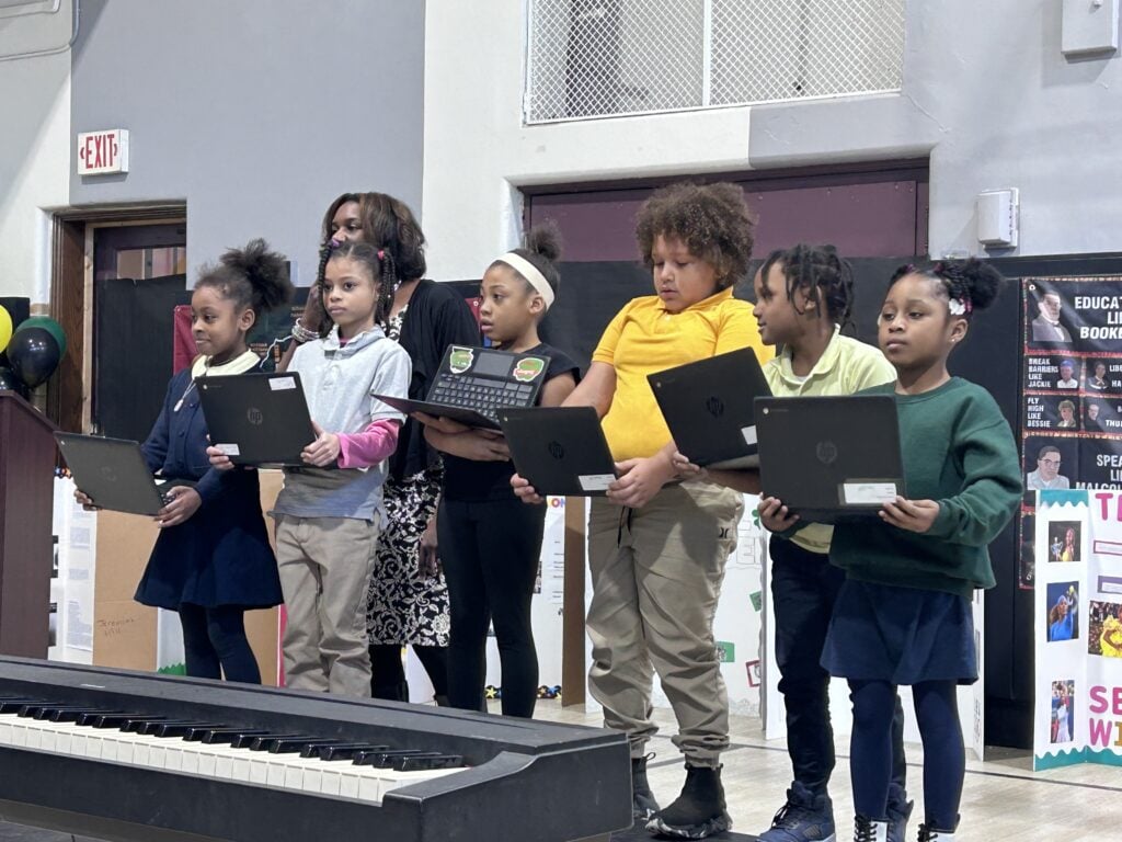 Children and an adult on stage, each child holding a laptop.