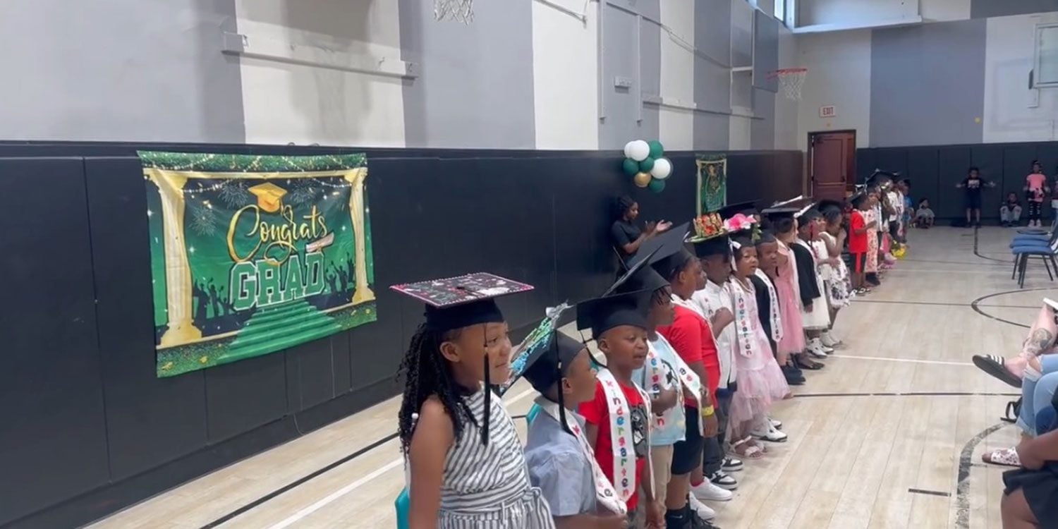 Children in graduation caps and sashes lined up in a gym.