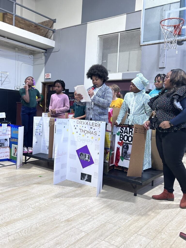 Children and an adult presenting projects in a school gym.