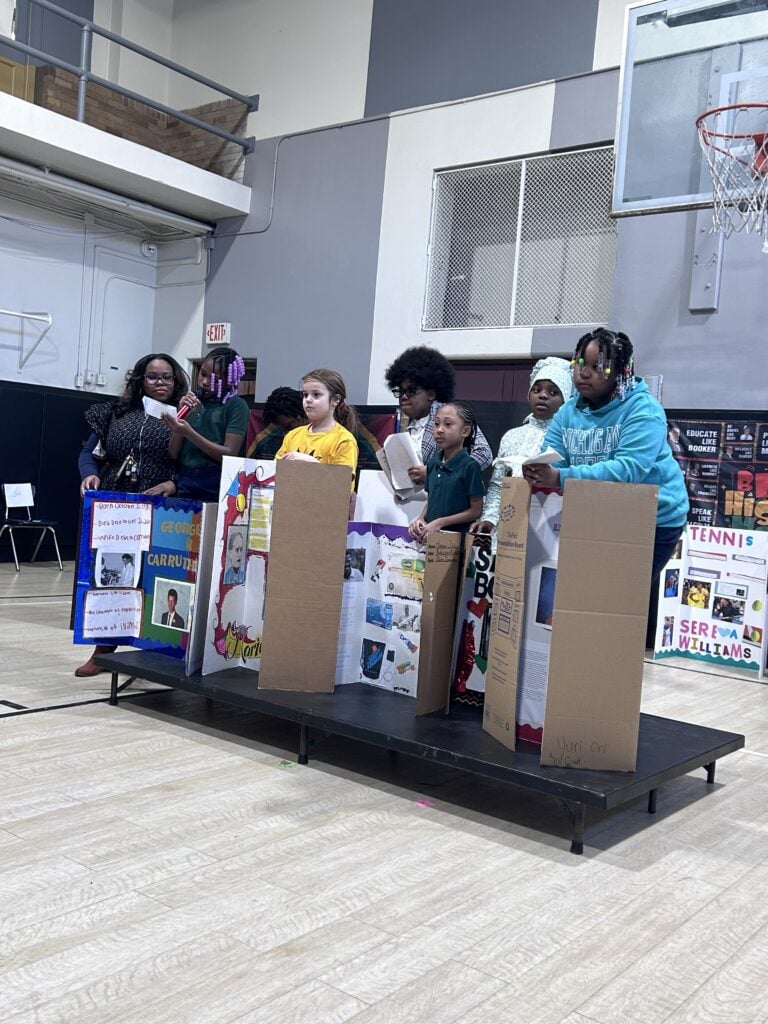 Children and an adult present science fair projects on a stage.