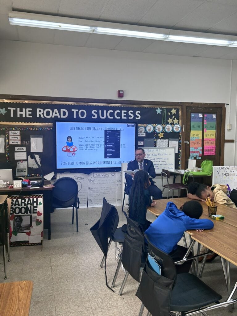Adult teaching children in a classroom with a large screen display.