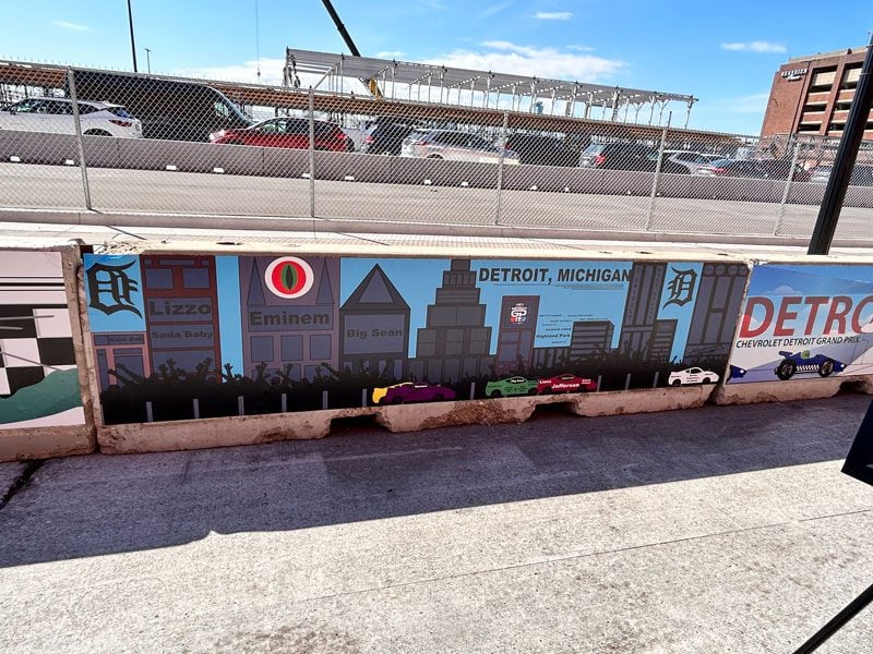 Mural on concrete barriers with Detroit cityscape, musicians' names, and race cars.