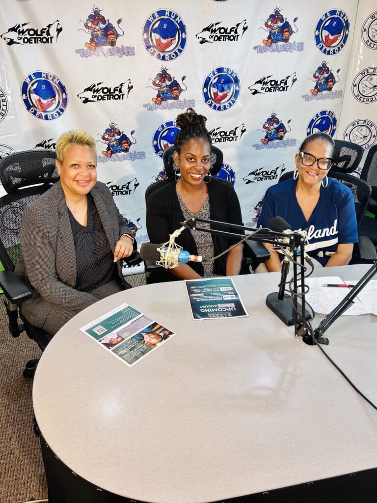 Three adults at a table with microphones in a radio studio.
