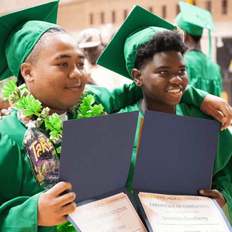 Two graduates in green caps and gowns holding certificates.